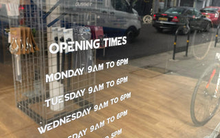 shop window with opening times painted on; faint view of inside shop, with bike parts and bikes; reflections of traffic in hornsey road
