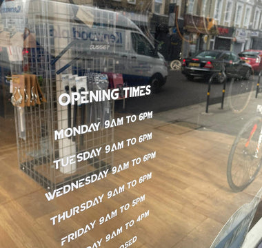 shop window with opening times painted on; faint view of inside shop, with bike parts and bikes; reflections of traffic in hornsey road